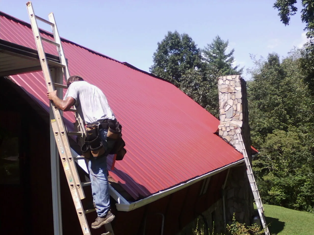 Roofer on a ladder beside a red metal roof and stone chimney — Soffit & Fascia Repair by Roofing Brothers in Mary D